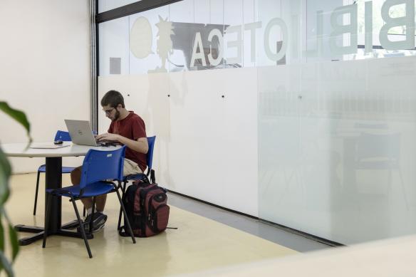 Foto de jovem branco usando camiseta vermelha e bermuda clara sentado em uma cadeira azul em frente a uma mesa redonda, utilizando um notebook. Há uma mochila encostada na cadeira. A cena ocorre em um espaço interno, ao lado de uma parede de vidro fosco com a palavra "BIBLIOTECA" escrita em grandes letras (vista ao contrário por estar do lado interno). Ao fundo, há um adesivo na parede com a silhueta de um personagem de desenho animado e um balão de fala.