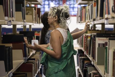 Foto de duas pessoas entre estantes de livros. A pessoa em primeiro plano tem cabelos cacheados e presos, usa óculos, brincos grandes de argola, uma regata branca e uma jaqueta verde caída nos ombros. Ela segura um papel com anotações enquanto busca um livro. Atrás, outra pessoa, de cabelos longos e escuros, também procura um livro segurando um papel. As estantes contêm livros organizados com etiquetas, e há uma janela ao fundo.