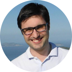 Foto de homem branco sorrindo. Ele tem cabelos curtos, lisos e pretos e usa óculos. Ele está com uma camisa de botões branca e ao fundo, uma paisagem de mar e céu azul.