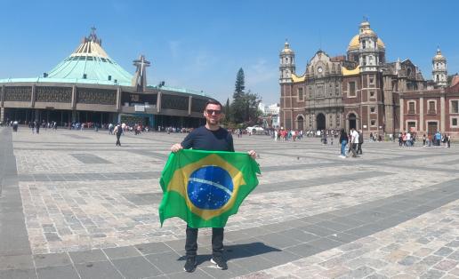 Foto de Wellington em frente à Basílica de Nossa Senhora de Guadalupe, na cidade do México. Wellington está de pé e segura uma bandeira do Brasil. É um homem branco, de cabelos castanhos, barba castanha e levemente grisalha, veste camiseta azul marinha, calça preta e tênis preto. Está no centro da imagem. Ao fundo e à esquerda, está a Basílica; um prédio arredondado, com telhado cônico nas cores verde água e branco, com uma cruz em sua fachada. À direita, está uma construção antiga com paredes cor de tijolos e  com torres em seus quatro cantos, uma abóbada amarela no centro, portas ovaladas e um relógio no centro de sua fachada. 