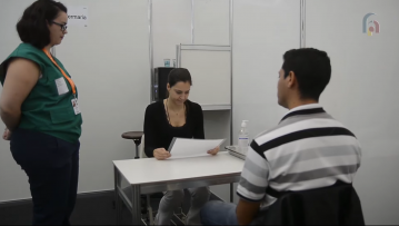 Foto de um atendimento simulado. Uma mulher branca vestida de preto está sentada. Ela se apoia em uma mesa para ler um papel. Um homem branco de cabelos curtos e escuros está sentado na frente dela e de costas para a câmera. Ao lado da mesa, uma mulher branca que usa óculos e tem cabelos escuros observa a cena.