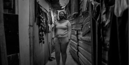 Foto em preto e branco de uma mulher em um corredor entre casas de uma comunidade. À sua direita, estão penduradas algumas roupas. A mulher usa máscara, veste blusa de manga longa e está com as mãos nos bolsos das calças.