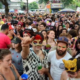 grupo de pessoas na rua, em bloco de carnaval. No centro da imagem, um jovem de máscara de carnaval olha diretamente para a câmera. Ao fundo, carro de som.