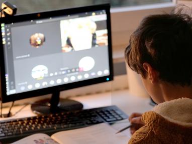 Foto colorida, em ambiente interno, criança em frente à tela do computador em chamada de vídeo