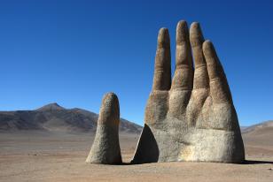 Foto colorida em ambiente externo, da escultura Mano del Desierto, que consiste em uma mão projetada em direção ao céu em meio ao deserto de Atacama.