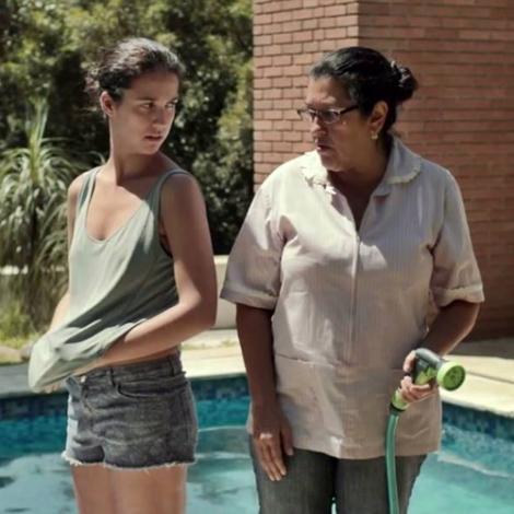 Cena do filme "Que horas ela volta?" onde as duas personagens principais se encaram na frente de uma piscina. Do lado esquerdo, a personagem Jéssica usa uma regata verde clara e um shorts jeans. Do lado direito, a personagem Val uma calça jeans e uma camisa grande e branca, ela também segura uma mangueira verde.