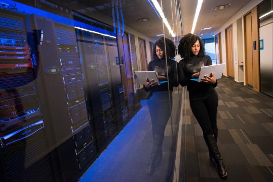 Foto de uma mulher jovem, negra, de cabelos crespos médios e escuros e vestida de preto segurando um notebook prata. Ela está em pé em um corredor e se apoia em um vidro escuro. Atrás do vidro, há muitas máquinas e equipamentos de armazenamento de dados iluminados por uma luz azul.