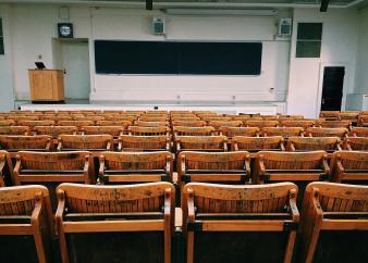 Foto colorida, em ambiente interno, de sala de aula com cadeiras de madeira e quadro preto