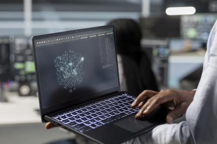 Foto de pessoa negra de perfil, segurando um notebook. Na tela escura, a visualização de dados na forma de um globo de pontos conectados em rede. Uma mão opera o mouse do teclado iluminado. Ao fundo, desfocado, mesas de trabalho e outros computadores.