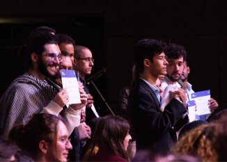 Cinco jovens, um ao lado do outro, estão de pé e seguram o certificado de conclusão de curso. Eles olham para frente e sorriem. Outro alunos, sentados, observam à frente