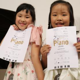 Na foto, duas meninas chinesas (Muyan Xiao e Letícia Guo Lin) seguram certificados e medalhas penduradas no pescoço. As duas têm cabelos pretos e lisos. Elas usam vestidos em tons claros, estampados com flores, e estão sorrindo, olhando para a câmera. A menina da esquerda tem um laço rosa claro no ombro do vestido e a menina da esquerda usa um penteado com trança.