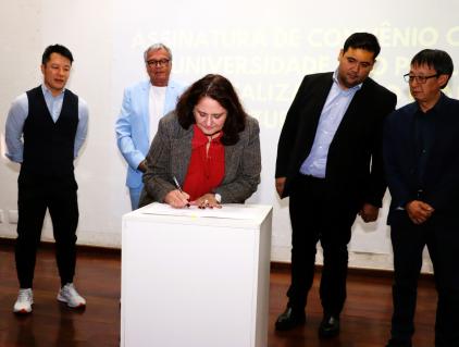 Foto de uma mulher parda, de cabelos de tamanho médio e castanhos, vestindo uma camisa vermelha e um blazer cinza. Elá está inclinada sobre um pódio branco assinando um documento. Atrás dela, quatro homens em trajes formais a observam. Ao fundo, um texto projetado, em letras amarelas, numa parede branca.
