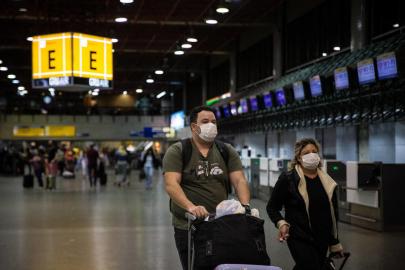 Foto tirada em área de check-in de um aeroporto. Um homem e uma mulher aparecem em primeiro plano usando máscaras e carregando malas.