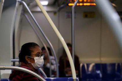 Foto de uma mulher de meia idade vestida de vermelho sentada em um vagão de metrô com poucas pessoas. A mulher usa máscara branca, brincos e óculos pretos. Ela tem seus cabelos curtos e escuros presos.