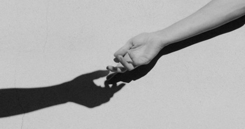 Foto em preto e branco de um braço estendido em frente a uma parede. Ao lado, tem-se uma sombra, também de um braço estendido, nessa mesma parede. Cria-se então uma ilusão de ótica como se a mão e a sombra se tocassem.
