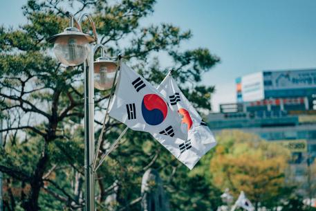Foto colorida em ambiente externo, da duas bandeiras da Coreia do Sul balançando, presas em um poste