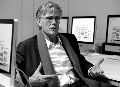 Foto em preto e branco de um senhor de cabelos curtos. Ele veste camisa, terno, cachecol e usa óculos. O homem está sentado em frente a alguns computadores.