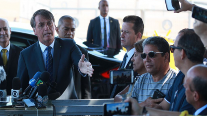 Foto colorida do presidente Bolsonaro discursando em meio a seguranças e apoiadores. No canto esquerdo, há microfones de diversas emissoras de televisão.