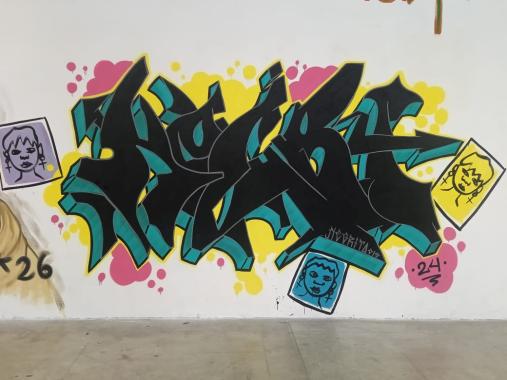 Foto de uma arte em Graffiti em uma parede branca, com caracteres de aglutinação típicos de pixação em um desenho tridimensional, em que as laterais das letras são verdes e a parte frontal delas é preta, no entorno da ilustração há detalhes em rosa e amarelo com formato de nuvens. Em volta da palavra graffitada, há também ilustrações em estilo retrato com ilustrações do mesmo rosto com brincos de cruz, nas cores roxa, azul e amarela
