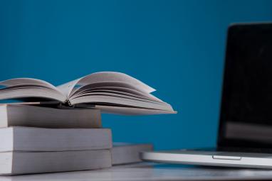 Foto colorida, em ambiente interno, pilha de livros ao lado de notebook aberto, em frente à parede azul