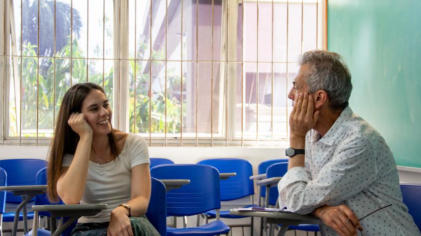 Foto de duas pessoas sentadas, uma mulher jovem e um senhor idoso, se olhando. Os dois encostam um de seus cotovelos na mesa e apoiam a cabeça na mão. Ela é branca, tem cabelos longos e lisos e veste uma camiseta branca. Ele é branco, tem cabelos curtos e grisalhos, usa um relógio preto e veste uma camisa branca. Ao redor deles, estão carteiras escolares azuis e, ao fundo, estão uma lousa e uma janela.