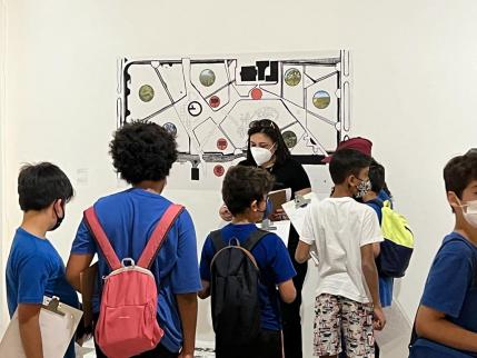 Sete estudantes, crianças do ensino fundamental 1, observam a professora - uma mulher branca de cabelos longos e negros e vestida com uma blusa e uma calça preta - ao fundo uma obra de arte na parede