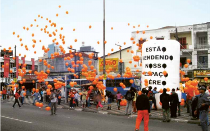 Foto tirada no meio da rua. Na calçada, há muitas pessoas e um inflável branco com o texto "Estão vendendo nosso espaço aéreo". O maior destaque são os diversos balões laranjas que aparecem voando na rua.