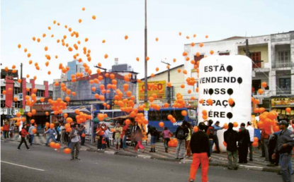 Foto tirada no meio da rua. Na calçada, há muitas pessoas e um inflável branco com o texto &quot;Estão vendendo nosso espaço aéreo&quot;. O maior destaque são os diversos balões laranjas que aparecem voando na rua.