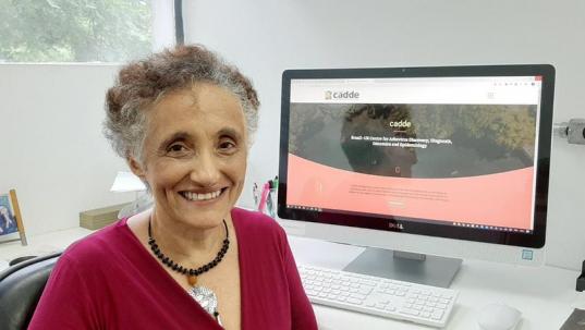 Foto de Ester Sabino, uma mulher sentada em frente a um computador sorrindo. Ela é branca, tem cabelos curtos e grisalhos, veste uma blusa vermelha e usa um colar artesanal. Atrás dela, há paredes brancas e alguns materiais de escritório sobre a mesa.
