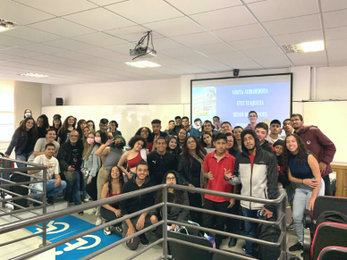 Grupo de cerca de 30 estudantes posam para a foto. Eles estão em um auditório, em frente da tela de projeção.