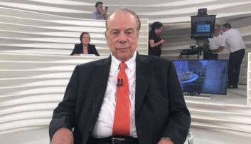 Foto de Ethevaldo Siqueira. Um homem branco, parcialmente calvo e grisalho. Na foto, ele está sentado em uma cadeira em um estúdio de TV. Veste um terno preto, camisa branca e gravata vermelha. Ao fundo, três pessoas, dois homens e duas mulheres, ajustam os equipamentos para gravação do programa de TV.