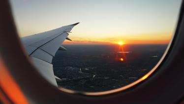 Foto colorida em ambiente interno, de uma janela de um avião com vista panorâmica para uma cidade, e, ao fundo, o pôr do sol.