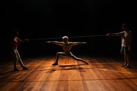 Foto de três pessoas em um palco. Ao centro há um homem que estica os braços e as pernas em uma pose que faz um ângulo reto com uma perna e estica a outra em uma inclinação acentuada. O centro do corpo do homem coincide com o ponto de cruzamento de três linhas retas desenhadas no palco. Um homem e uma mulher estão de pé nas extremidades da imagem. Cada um deles segura a ponta de um cordão que passa por cima da cabeça do homem ao centro. O fundo do palco é preto e a luz vem de cima e marca apenas a silhueta dos artistas.