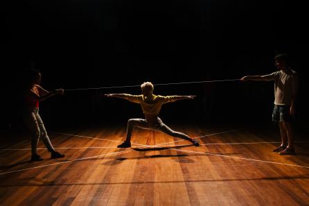 Foto de três pessoas em um palco. Ao centro há um homem que estica os braços e as pernas em uma pose que faz um ângulo reto com uma perna e estica a outra em uma inclinação acentuada. O centro do corpo do homem coincide com o ponto de cruzamento de três linhas retas desenhadas no palco. Um homem e uma mulher estão de pé nas extremidades da imagem. Cada um deles segura a ponta de um cordão que passa por cima da cabeça do homem ao centro. O fundo do palco é preto e a luz vem de cima e marca apenas a silhueta dos artistas. 