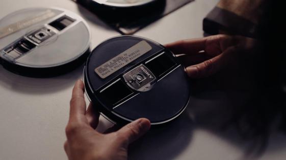 Foto de fitas de filme sobre uma mesa branca. No centro, duas mãos brancas seguram um rolo de fita filmográfica com uma etiqueta que diz She has a beard, by Norma Bahia e Rita Moreira, black and white, 30 minutes.