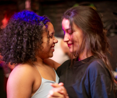 Descrição acessível: foto de duas mulheres dançando próximas, uma de frente para a outra e de perfil com fundo desfocado. A da esquerda é negra, de cabelos cacheados curtos, está com uma faixa na cabeça, blusinha acinzentada e está sorrindo. A da direita é branca, de cabelos castanhos, blusa azul escura e está sorrindo.