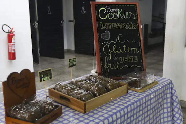 Foto de uma banca de produtos. Em primeiro plano, há uma mesa coberta com uma toalha xadrez azul e branco. Sobre a mesa há três suportes com cookies embalados. Da esquerda para a direita há duas caixas de cookies e placas com os dizeres “glúten e leite free” e “choco chip”. À direita há uma mini-lousa escolar com os dizeres “Bono Cookies, integrais e glúten e leite free”, tendo à frente o terceiro suporte de cookies com uma tampa acrílica transparente. A mesa está em uma sala com paredes brancas, piso cinza e, ao fundo, há um extintor e portas para banheiros.