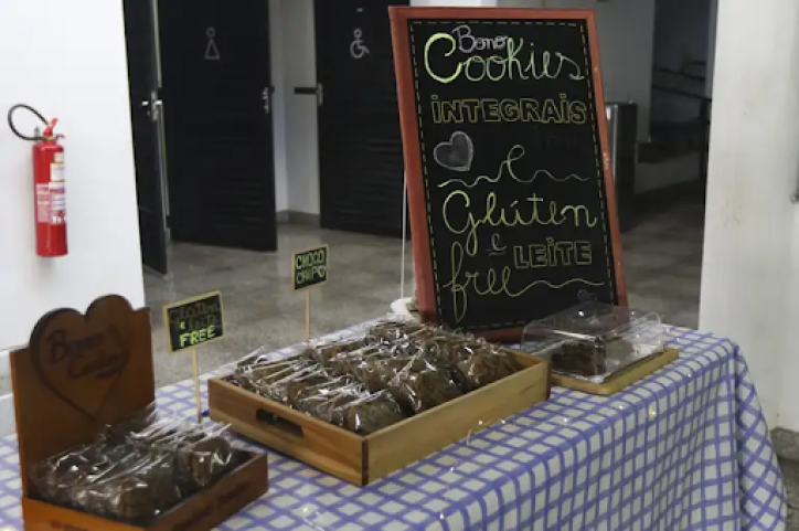 Foto de uma banca de produtos. Em primeiro plano, há uma mesa coberta com uma toalha xadrez azul e branco. Sobre a mesa há três suportes com cookies embalados. Da esquerda para a direita há duas  caixas de cookies e  placas com os dizeres “glúten e leite free” e “choco chip”. À direita há uma  mini-lousa escolar com os dizeres “Bono Cookies, integrais e glúten e leite free”, tendo à frente o terceiro suporte de cookies com uma tampa acrílica transparente. A mesa está em uma sala com paredes brancas, piso cinza e, ao fundo, há um extintor e portas para banheiros.