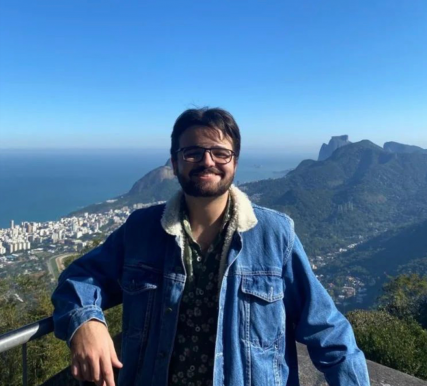 Foto de um rapaz sorrindo em uma paisagem do Rio de Janeiro. Ele é branco, tem cabelos lisos escuros, usa barba e óculos. O jovem veste uma camisa verde e uma jaqueta jeans.