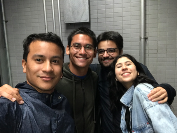 Selfie de quatro jovens sorrindo. São três rapazes e uma moça. Todos vestem blusas de frio e se abraçam.