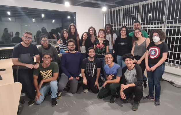 Foto de diversos jovens sorrindo para a câmera. Os estudantes se juntam para posar em uma foto na sala de apoio do estúdio do CJE. Pedro está agachado no centro do grupo. Ele veste uma blusa de manga longa roxa e uma calça cinza.