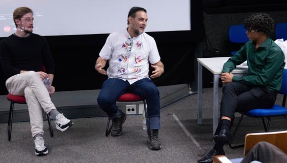Foto de três homens falando em um auditório. Na esquerda, um homem branco de cabelos e olhos claros vestindo blusa preta e calça clara, no meio, um homem branco de camisa branca com bordados coloridos e calça azul, na direita, homem negro de camisa verde e calça azul. Eles estão sentados em cadeiras com estofados vermelhos ou azuis, o piso tem carpete cinza e atrás deles há um pequeno palco e uma tela de projeção.
