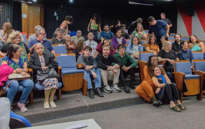 Foto de pouco mais de 30 pessoas em plateia de auditório com cadeiras de madeira e estofado azul e carpete cinza. Na frente, há pessoas sentadas e algumas pessoas, em fileiras mais distantes, estão de pé. No fundo há paredes pretas e uma porta de madeira.