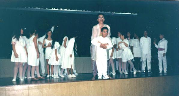 Foto de Luiz de Godoy em uma apresentação dos Pequenos Cantores de Mogi das Cruzes, grupo de coral do local. Luiz é uma criança negra, veste uma blusa de manga longa branca, calça branca e tênis branco. Atrás dele, o abraçando, está a regente do coral, uma mulher negra, de cabelos escuros, usando roupas brancas e óculos. Ao fundo, estão oito crianças do lado esquerdo e oito crianças do lado direito. Todas estão vestidas de branco. Todos os integrantes do coral estão num palco de madeira, o fundo é escuro.