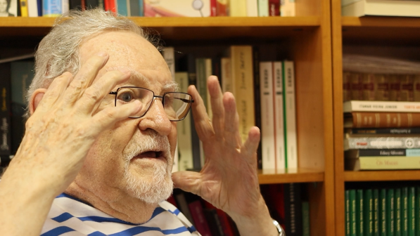 Foto de homem branco de olhos escuros, cabelos e barba curtos e grisalhos falando e gesticulando com as mãos próximas à cabeça. Ele usa óculos e uma camisa branca com listras azuis. Atrás dele, há estantes com livros.