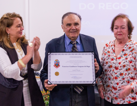 Foto de três pessoas em pé, lado a lado. No centro, um homem branco, idoso, vestindo terno escuro, camisa azul e gravata listrada, segurando e exibindo um certificado. À esquerda, uma mulher branca de cabelos loiros e longos, vestida com uma roupa preta e branca, está batendo palmas. À direita, há outra mulher branca, idosa, de cabelos castanhos curtos, vestindo uma blusa florida. Ao fundo, tela de projeção exibindo texto em azul e branco.