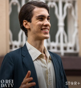 Foto do estudante de Publicidade e Propaganda e ator, Guilherme Reis, durante gravação para o seriado &quot;O Rei da TV&quot;. Guilherme está em primeiro plano, sorrindo, caracterizado como seu personagem. Ele é branco e tem cabelo castanho, usa um blazer cinza escuro e uma camisa bege. Ao fundo da imagem, aparecem as grades de uma casa antiga.