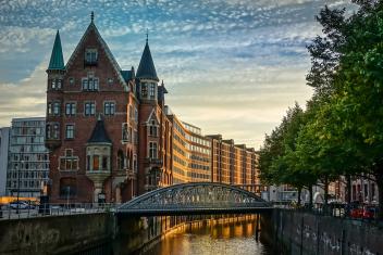 Foto colorida em ambiente externo, de uma locação urbana na cidade de Hamburgo, na Alemanha, com um céu claro e azul, prédios típicos da arquitetura alemã e árvores.