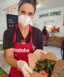 Foto de uma mulher segurando uma pequena caixa de papelão com comida dentro. Ela é branca, tem olhos escuros, cabelos presos, usa máscara e luvas descartáveis e veste uma camiseta preta e um avental vermelho. Ao fundo, há um cartaz na parede branca com o texto “juntos somos” em preto.