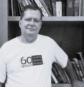 Foto em preto e branco. Um homem branco, de cabelos curtos e ligeiramente grisalhos, posa para uma fotografia. Ele veste uma camiseta branca, com uma estampa em preto com o texto 60 anos Conservatório de Tatui. Atrás dele, há um estante com livros. Ele apoia uma das mãos na estante.